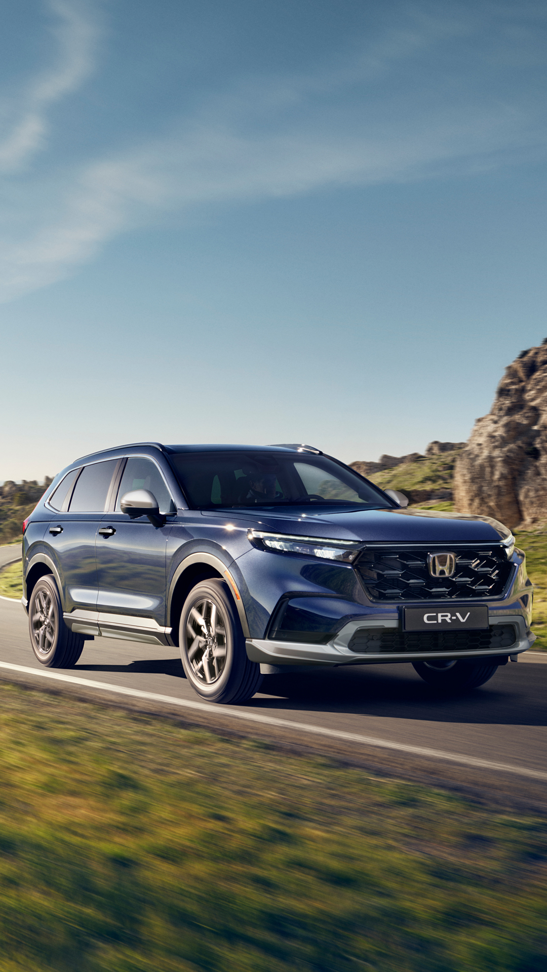 Ein dunkelblauer Honda CR-V fährt eine kurvenreiche Bergstrasse entlang, umgeben von üppigem grünem Gras und felsigem Gelände. Die Landschaft zeichnet sich durch sanfte Hügel und ferne Berge unter einem klaren blauen Himmel mit vereinzelten Wolken aus.
