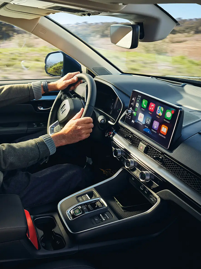 Interior view of a modern car with a driver seated behind the wheel, showing the dashboard, steering wheel, center console, and a wide touchscreen display with colorful app icons, while countryside scenery passes by outside the windows.