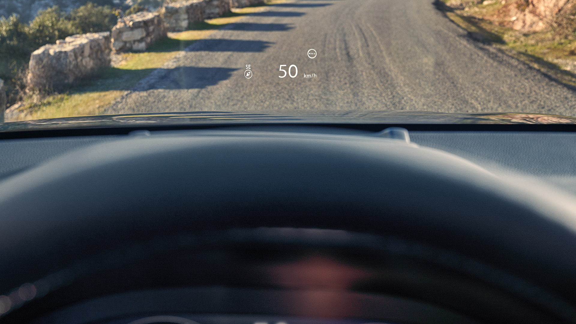 A first-person view from a driver's seat showing a Head-Up Display (HUD) projecting "50 km/h" and a compass onto the windshield above a gravel road.
