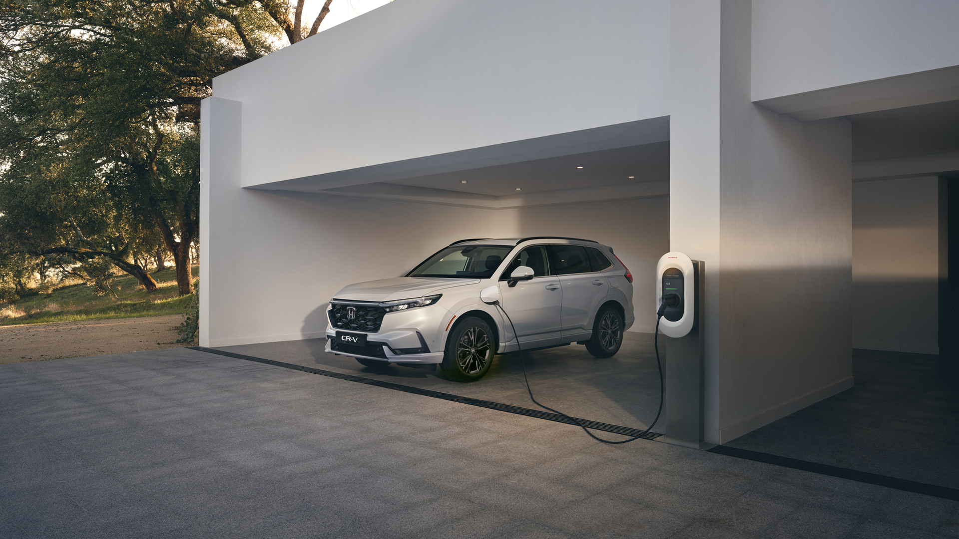 Ein weisser Honda CR-V Plug-in-Hybrid ist in einem minimalistischen weissen Carport geparkt und mit einem wandmontierten Elektrofahrzeug-Ladegerät verbunden.