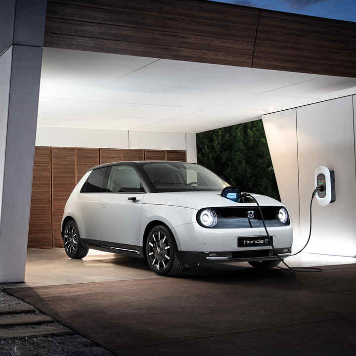 Honda e, 3/4-Frontansicht, ladend in Einfahrt geparkt, während Besitzer das Haus betritt.