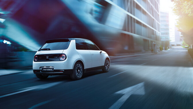 Honda e, 3/4-Frontansicht, beim Fahren durch eine elektrifizierte Stadt.