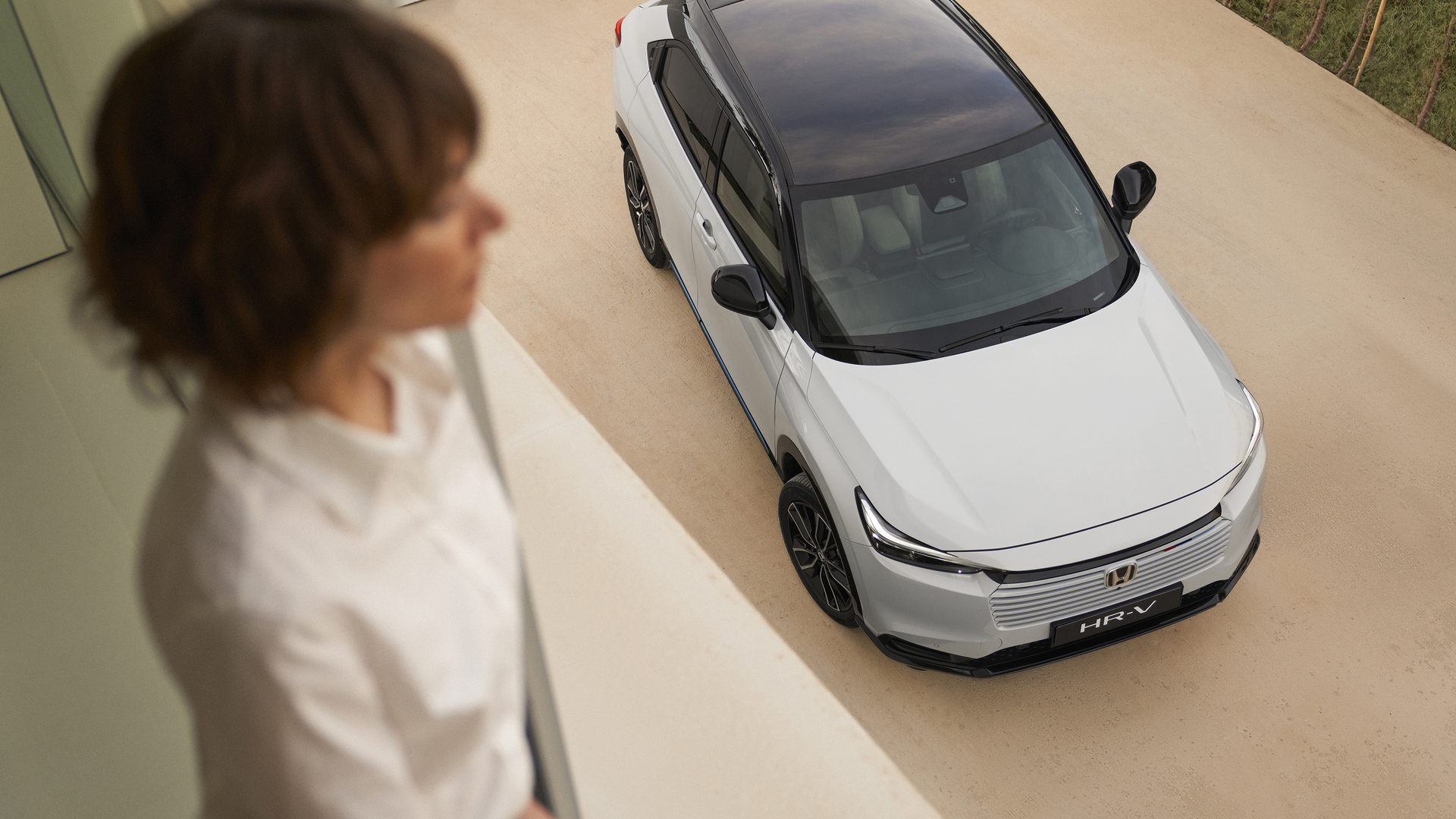 Blick von oben auf einen weißen Honda HR-V mit schwarzem Dach, der auf einer hellen Einfahrt geparkt ist, während im Vordergrund eine nicht deutlich erkennbare Person steht und auf das Fahrzeug hinunterblickt.
