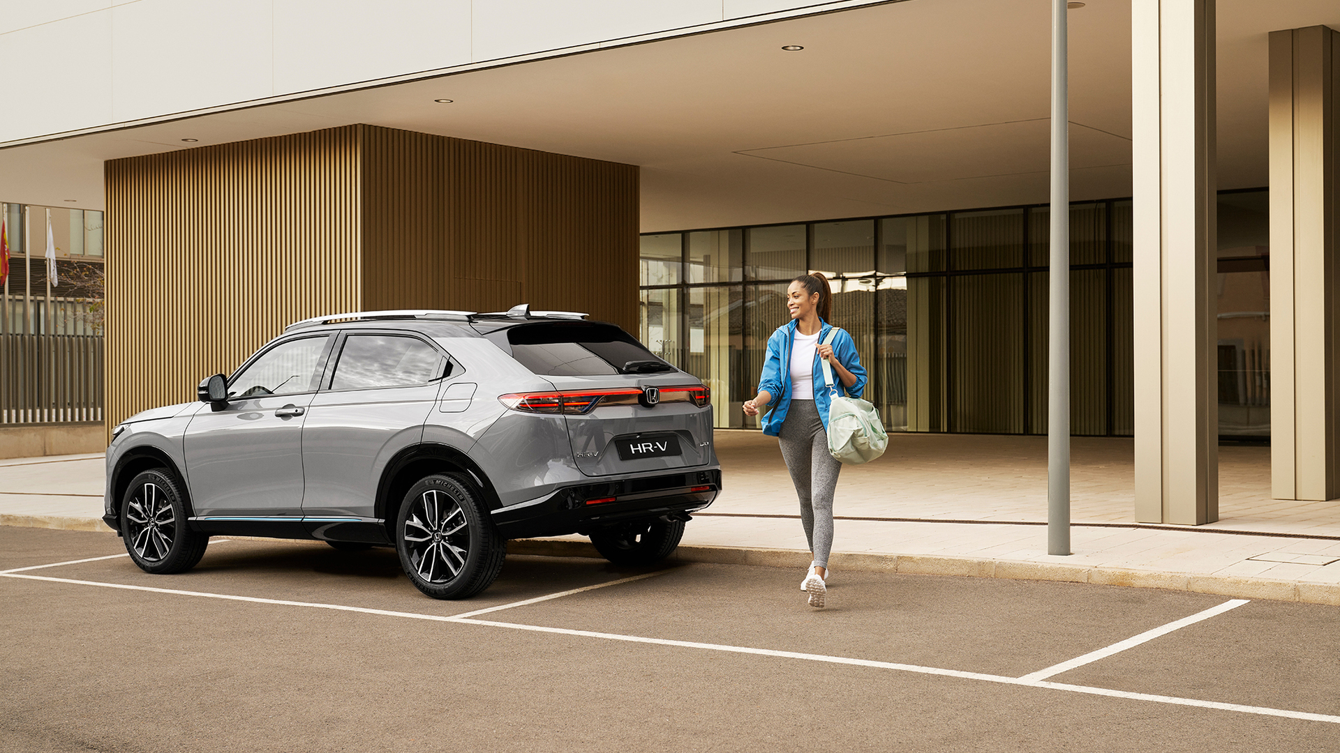 Ein silberner Honda HR-V steht vor einem modernen Gebäude – eine Frau mit einer Tasche geht daran vorbei, was auf den alltäglichen Einsatz in der Stadt und den damit verbundenen Komfort hinweist.