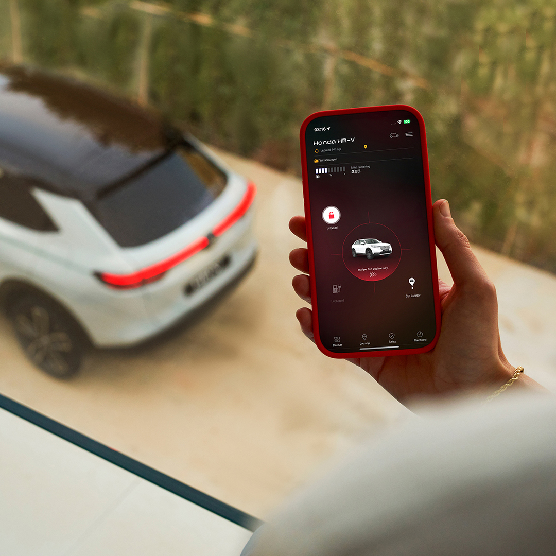 Eine Person hält ein Smartphone, auf dem eine App für Fahrzeugladeeinstellungen angezeigt wird, während im Hintergrund ein weißer SUV geparkt ist.