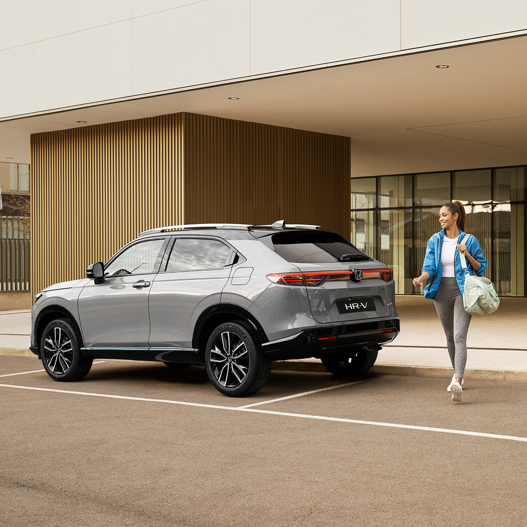 Ein silberner Honda HR-V steht vor einem modernen Gebäude – eine Frau mit einer Tasche geht daran vorbei, was auf den alltäglichen Einsatz in der Stadt und den damit verbundenen Komfort hinweist.