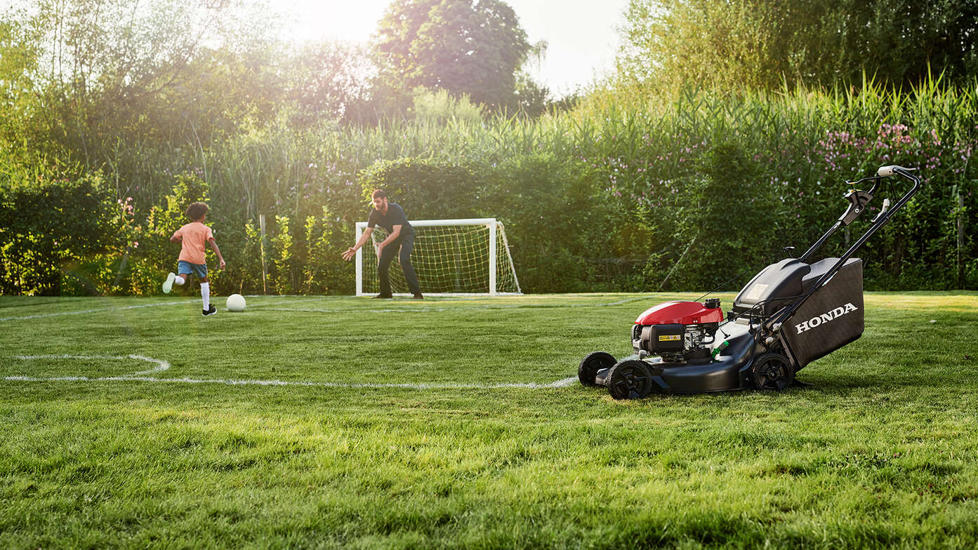 Mann und Kind beim Fussballspielen neben einem Honda HRN im Garten, Seitenansicht 