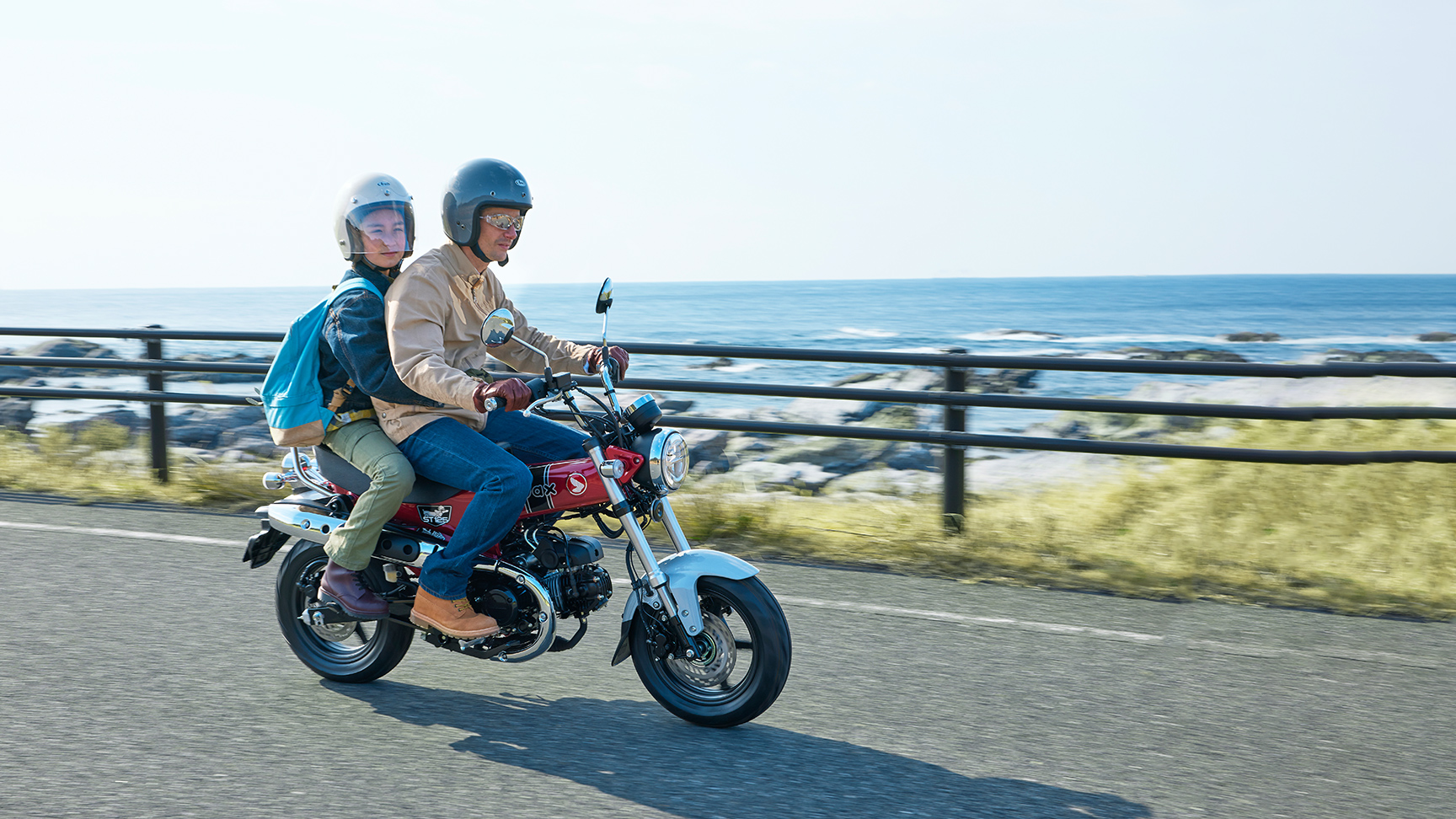 Zwei Personen mit Helmen fahren auf einem roten 125-ccm-MiniMotorrad eine Küstenstraße entlang; hinter einer Leitplanke sind Meereswellen und eine felsige Küste zu sehen.