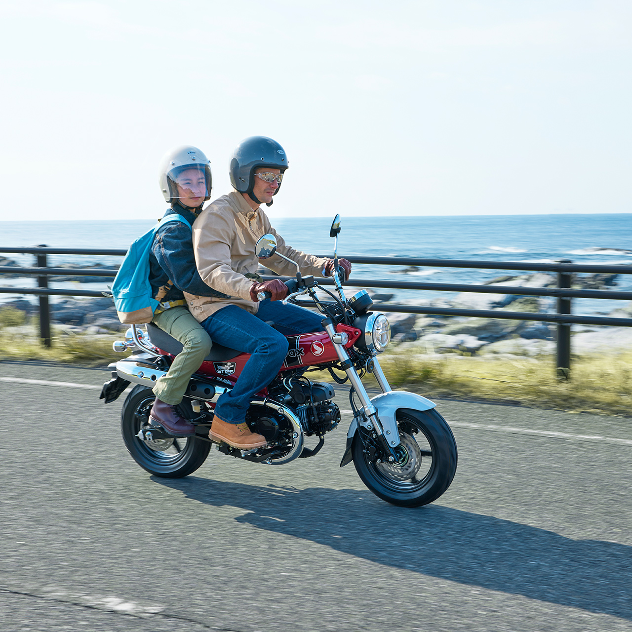 Zwei Personen mit Helmen fahren auf einem roten 125-ccm-MiniMotorrad eine Küstenstraße entlang; hinter einer Leitplanke sind Meereswellen und eine felsige Küste zu sehen.