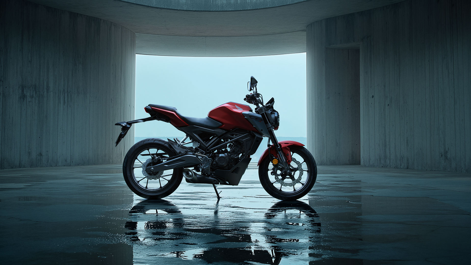 Ein rotes 125 ccm Mini-Motorrad, das an einer Strandpromenade neben einer Betonmauer geparkt ist, daneben liegen Helme und eine Tasche, im Hintergrund sind der Strand, das Meer und zwei Personen zu sehen.