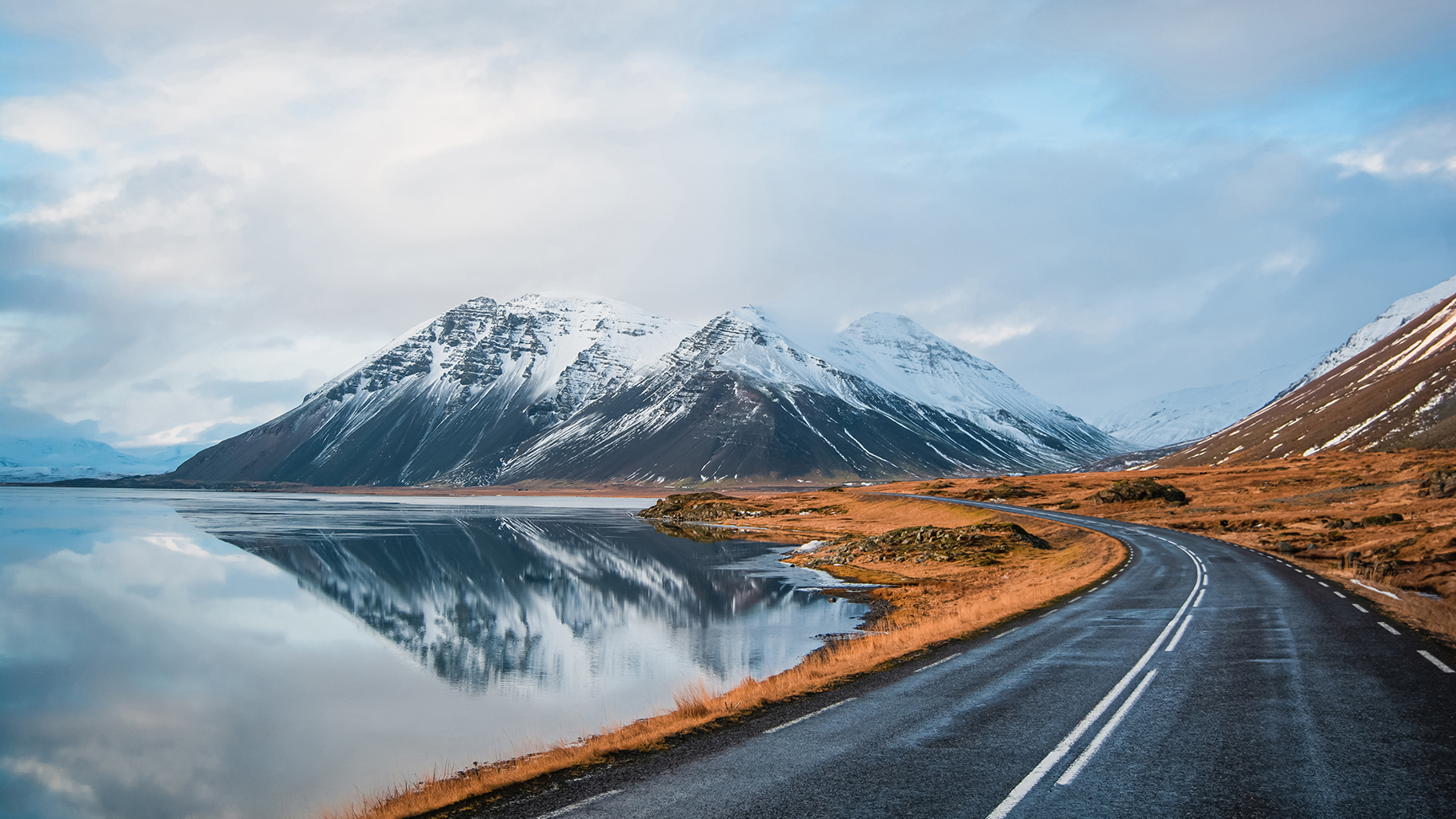 Panoramaansicht einer Straße im Winter, die entlang eines Seeufers zu Vulkanen führt. Spiegelbild hoher schneebedeckter Gipfel auf einer Wasseroberfläche. Blick aus der Fahrerperspektive auf die Ringstraße, Island.