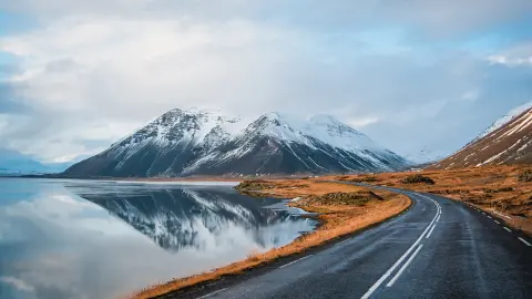 Panoramaansicht einer Straße im Winter, die entlang eines Seeufers zu Vulkanen führt. Spiegelbild hoher schneebedeckter Gipfel auf einer Wasseroberfläche. Blick aus der Fahrerperspektive auf die Ringstraße, Island.