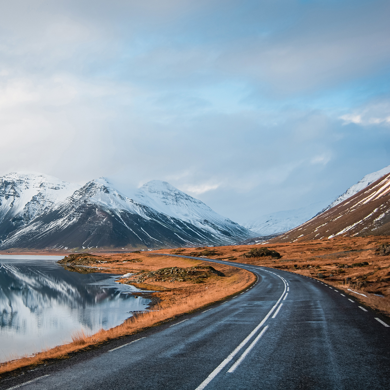 Panoramaansicht einer Straße im Winter, die entlang eines Seeufers zu Vulkanen führt. Spiegelbild hoher schneebedeckter Gipfel auf einer Wasseroberfläche. Blick aus der Fahrerperspektive auf die Ringstraße, Island.
