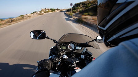 Blick auf das Motorrad-Cockpit, die Holme und die davor liegende Straße über die Schulter eines Fahrers hinweg.