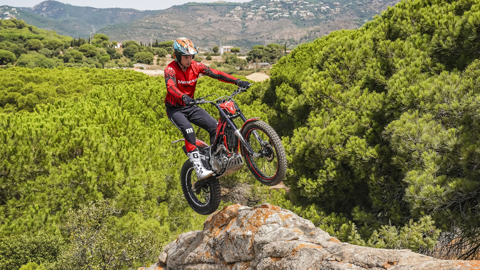 Ein Motorradfahrer springt mit einem roten Motorrad vom Typ Montesa Cota 4RT 260R von einem Felsvorsprung aus über eine malerische Kiefernwaldlandschaft.