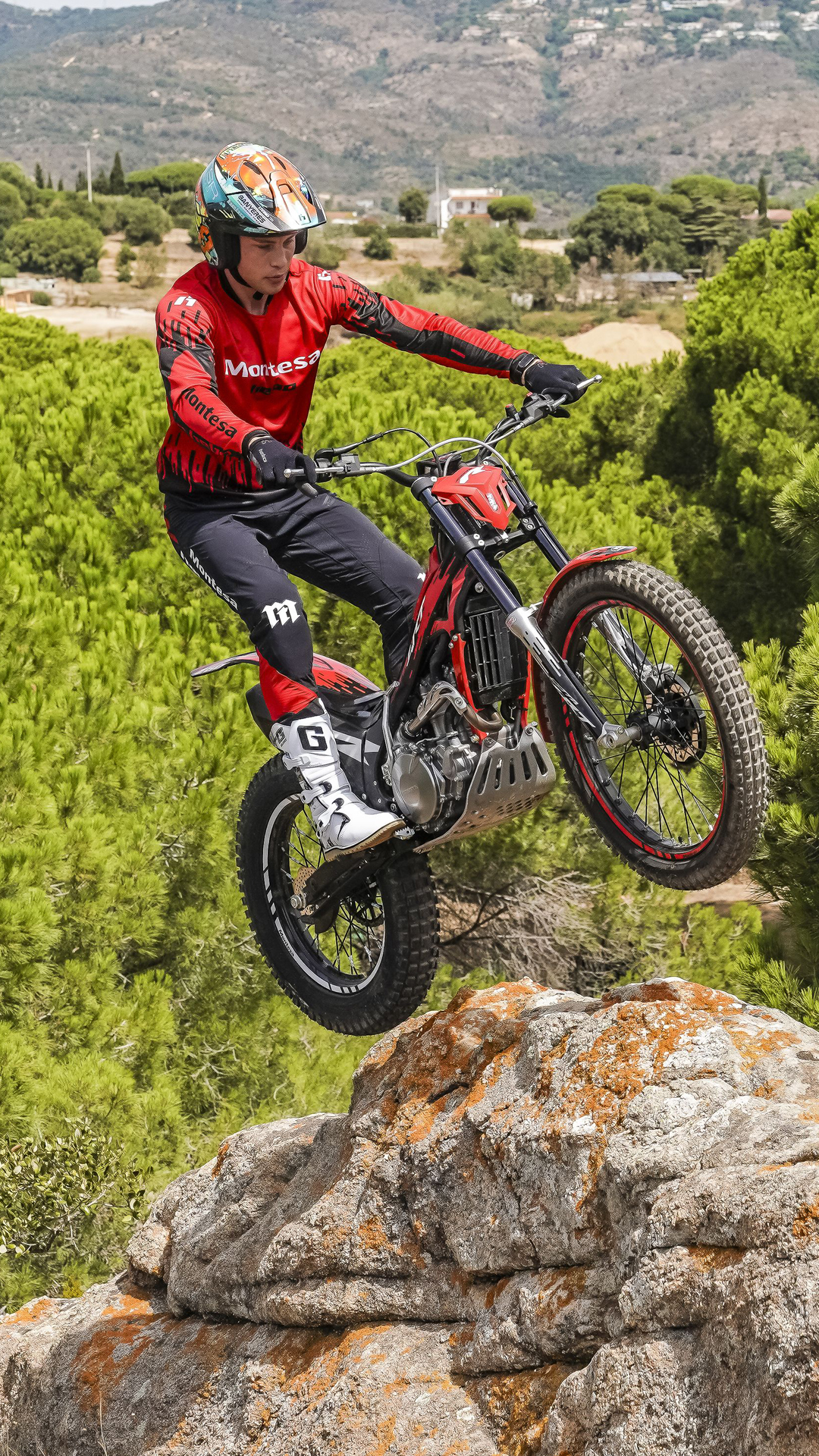 Ein Motorradfahrer springt mit einem roten Motorrad vom Typ Montesa Cota 4RT 260R von einem Felsvorsprung aus über eine malerische Kiefernwaldlandschaft.