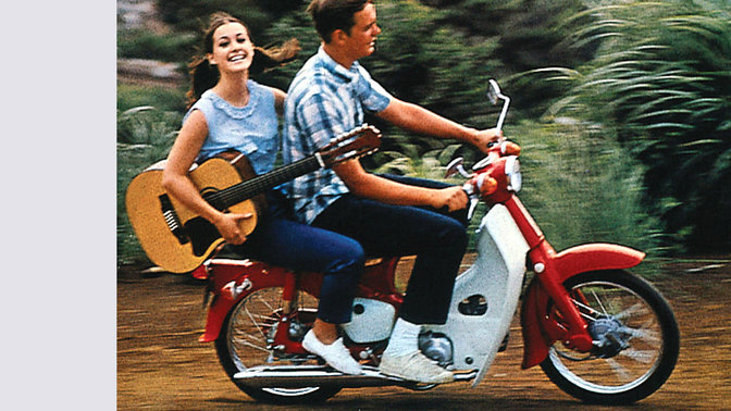 Honda Cub mit Fahrer und Beifahrerin mit Gitarre. Auf einer unbefestigten Strasse. Aufnahme von rechts.