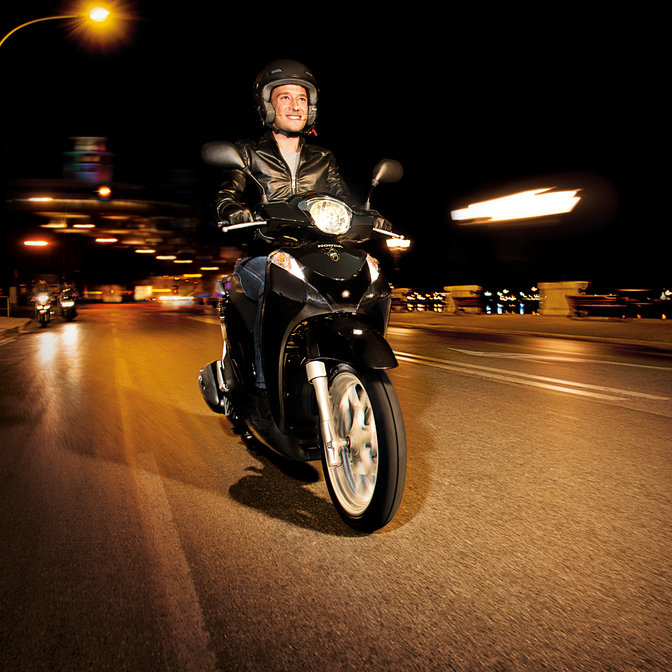 3/4-Frontansicht eines schwarzen Honda SH Mode-Motorrollers mit Fahrer. Nach rechts weisend (auf der Straße).