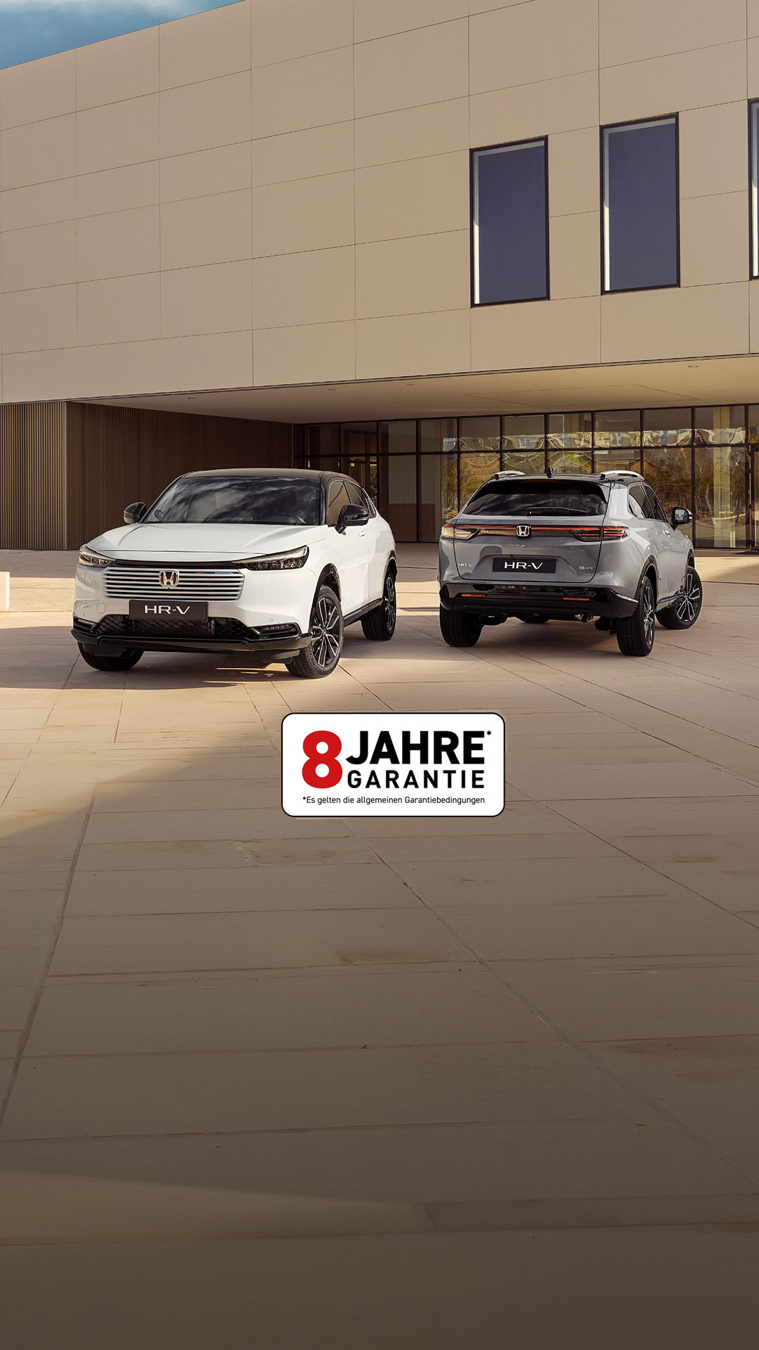 Zwei Honda HR-V SUVs, die vor einem modernen Gebäude geparkt sind, aus der Vorder- und Rückansicht – einer in Weiß, der nach vorne zeigt, und einer in Silber, der mit der Rückseite zum Betrachter zeigt.