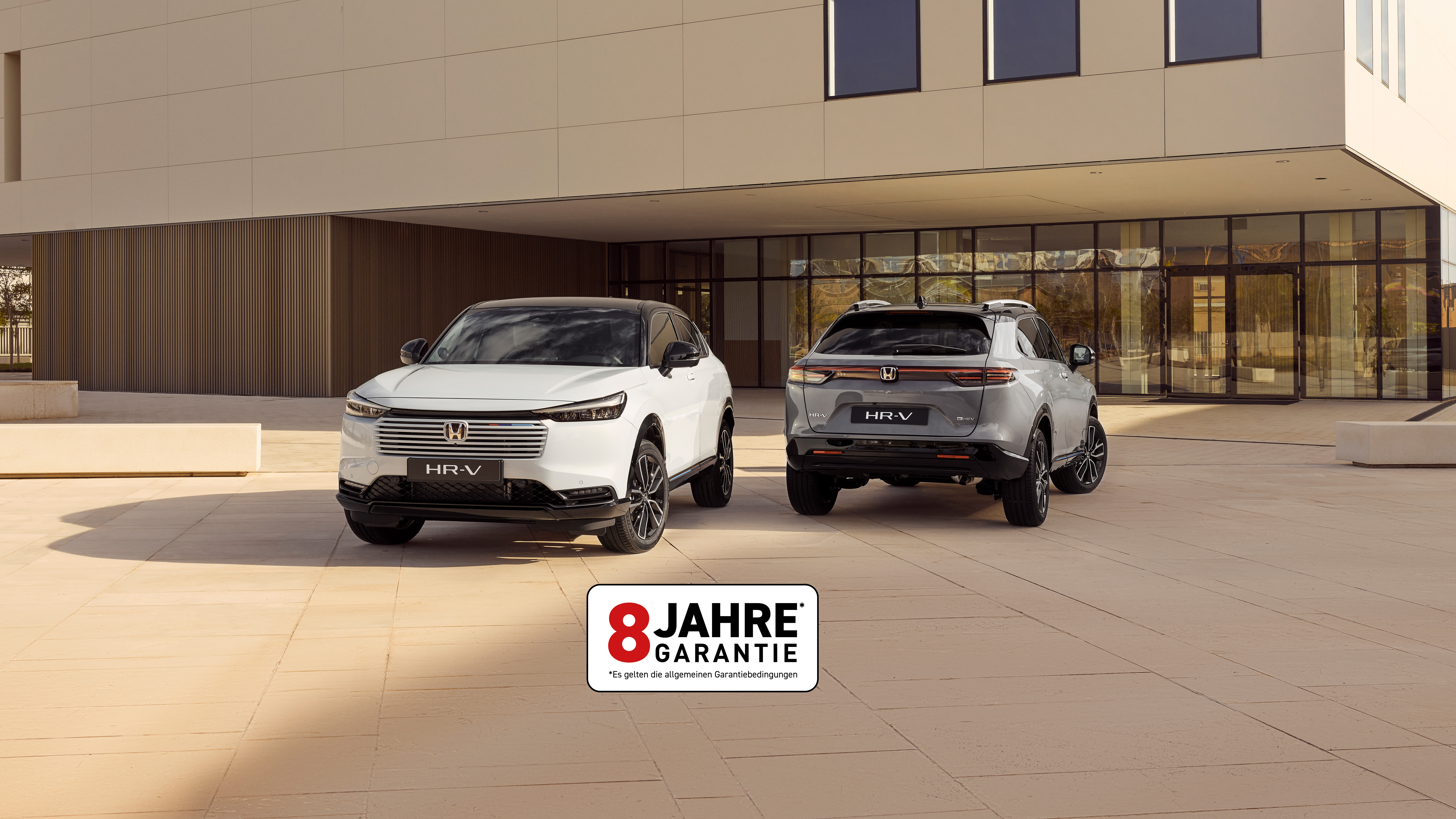 Zwei Honda HR-V SUVs, die vor einem modernen Gebäude geparkt sind, aus der Vorder- und Rückansicht – einer in Weiß, der nach vorne zeigt, und einer in Silber, der mit der Rückseite zum Betrachter zeigt.