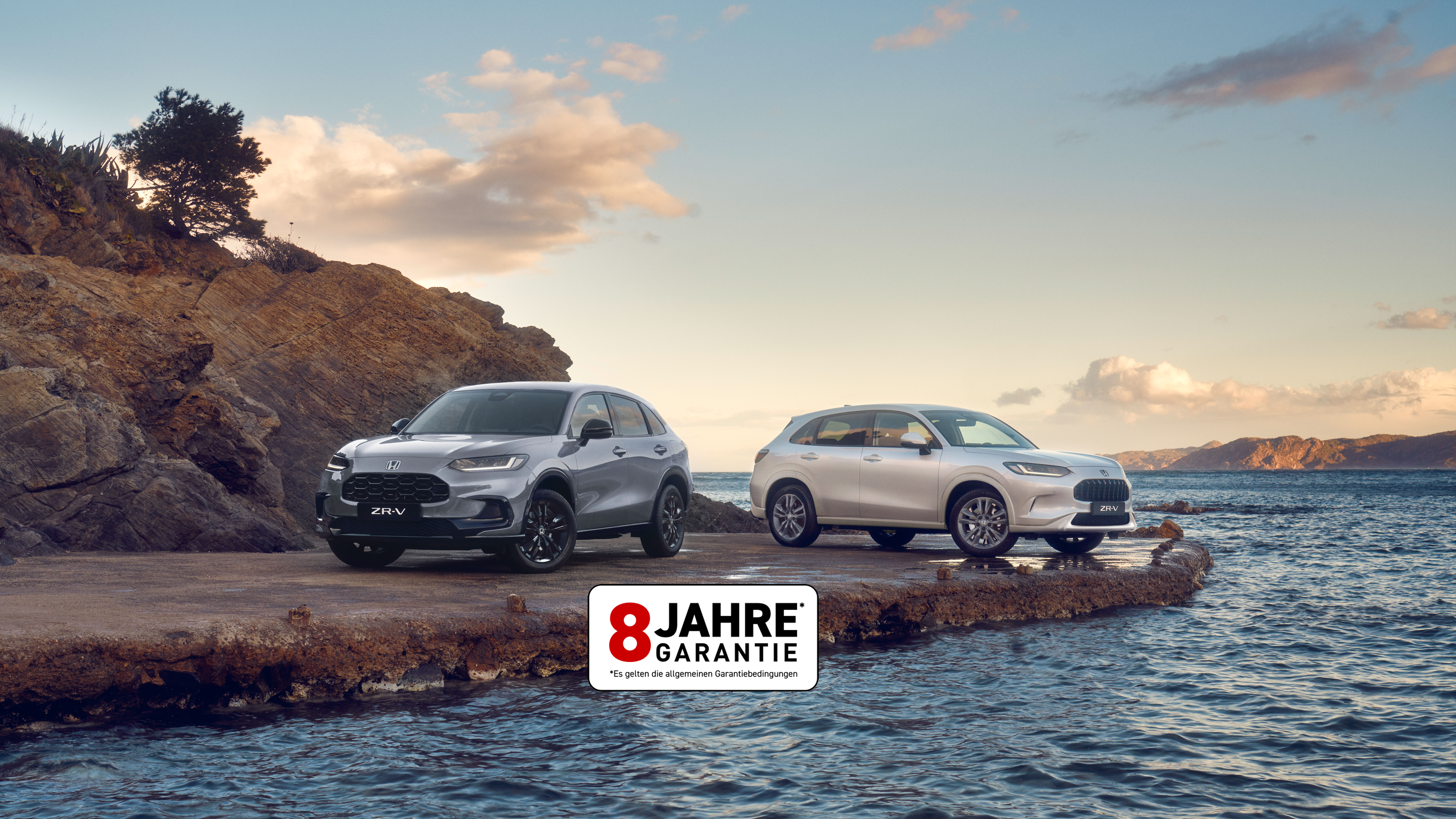 Two Honda ZR-V SUVs, one blue and one white, parked on a rocky coastal outcrop with water and hills in the background under a partly cloudy sky.