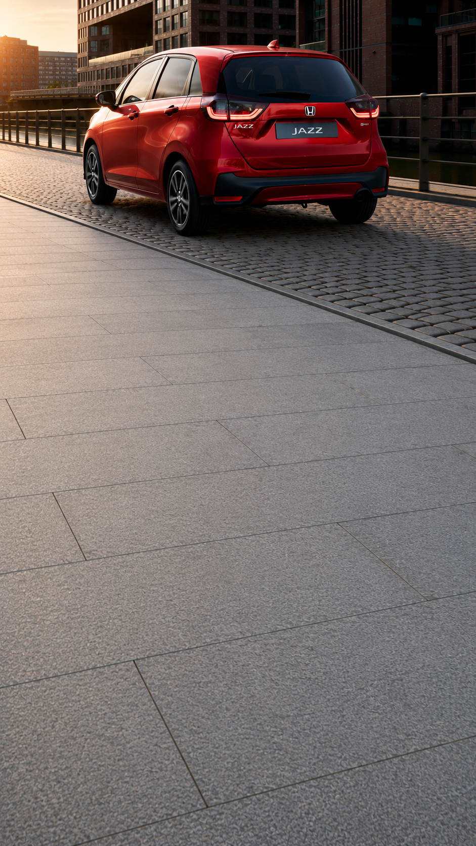 Honda Jazz in Premium Crystal Red Metallic fährt auf einer Stadtstrasse mit modernen Bürogebäuden im Hintergrund.